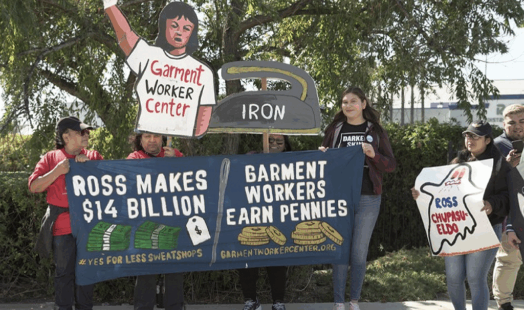 A small group gathers at a protest holding colorful homemade signs. One is the body of a woman wearing a t-shirt reading "Garment Worker Center." Another is an iron (for ironing clothes). A third holds a sign with a ghost that reads "Ross Chupasueldo." Two hold a banner reading "Ross makes $14 billion -- Garment Workers Earn Pennies"
